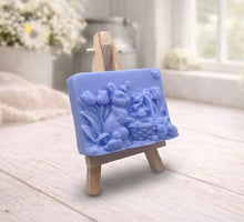 Load image into Gallery viewer, A rectangular handmade vegan soap in a sophisticated slate-grey tone, featuring a highly detailed relief scene of an Easter bunny standing next to a basket of eggs, surrounded by blooming tulips and small birds, displayed on a wooden easel.