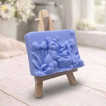 Load image into Gallery viewer, A rectangular handmade vegan soap in a sophisticated slate-grey tone, featuring a highly detailed relief scene of an Easter bunny standing next to a basket of eggs, surrounded by blooming tulips and small birds, displayed on a wooden easel.