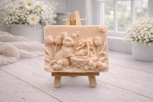 Load image into Gallery viewer, A rectangular handmade vegan soap in a sophisticated tone, featuring a highly detailed relief scene of an Easter bunny standing next to a basket of eggs, surrounded by blooming tulips and small birds, displayed on a wooden easel.