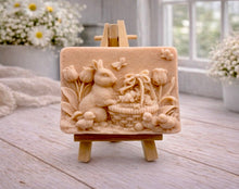 Load image into Gallery viewer, A rectangular handmade vegan soap in a sophisticated tone, featuring a highly detailed relief scene of an Easter bunny standing next to a basket of eggs, surrounded by blooming tulips and small birds, displayed on a wooden easel.