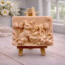 Load image into Gallery viewer, A rectangular handmade vegan soap in a sophisticated tone, featuring a highly detailed relief scene of an Easter bunny standing next to a basket of eggs, surrounded by blooming tulips and small birds, displayed on a wooden easel.