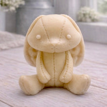 Load image into Gallery viewer, A handmade vegan soap sculpted in the shape of a sitting floppy-eared bunny rabbit with visible stitched seam details like a plush toy, in a soft yellow tone, displayed on a light wooden surface.