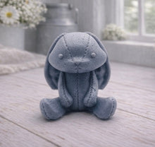 Load image into Gallery viewer, A handmade vegan soap sculpted in the shape of a sitting floppy-eared bunny rabbit with visible stitched seam details like a plush toy, in a soft grey tone, displayed on a light wooden surface.