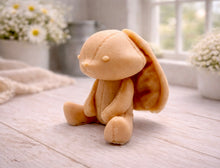 Load image into Gallery viewer, A handmade vegan soap sculpted in the shape of a sitting floppy-eared bunny rabbit with visible stitched seam details like a plush toy, in a soft peach-orange tone, displayed on a light wooden surface.
