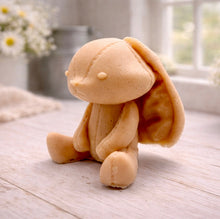 Load image into Gallery viewer, A handmade vegan soap sculpted in the shape of a sitting floppy-eared bunny rabbit with visible stitched seam details like a plush toy, in a soft peach-orange tone, displayed on a light wooden surface.