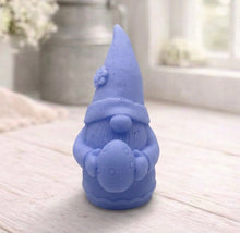 Load image into Gallery viewer, handmade vegan soap in the shape of a sitting gnome holding a decorated Easter egg, featuring a tall pointed hat with a flower detail and a textured beard, displayed on a neutral cloth in a bright spring setting.