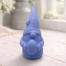 Load image into Gallery viewer, handmade vegan soap in the shape of a sitting gnome holding a decorated Easter egg, featuring a tall pointed hat with a flower detail and a textured beard, displayed on a neutral cloth in a bright spring setting.