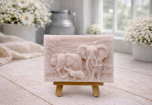 Load image into Gallery viewer, A rectangular handmade vegan soap, featuring a detailed relief carving of an elephant family—two adults and a small calf—walking through a textured savannah landscape, displayed on a miniature wooden easel.