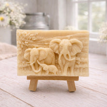 Load image into Gallery viewer, A rectangular handmade vegan soap in a warm yellow tone, featuring a detailed relief carving of an elephant family—two adults and a small calf—walking through a textured savannah landscape, displayed on a miniature wooden easel.