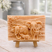 Load image into Gallery viewer, A rectangular handmade vegan soap in a warm terracotta tone, featuring a detailed relief carving of an elephant family—two adults and a small calf—walking through a textured savannah landscape, displayed on a miniature wooden easel.