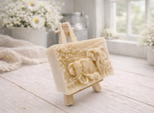 Load image into Gallery viewer, A rectangular handmade vegan soap in a warm yellow tone, featuring a detailed relief carving of an elephant family—two adults and a small calf—walking through a textured savannah landscape, displayed on a miniature wooden easel.
