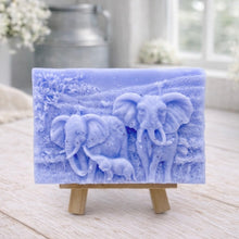 Load image into Gallery viewer, A rectangular handmade vegan soap, featuring a detailed relief carving of an elephant family—two adults and a small calf—walking through a textured savannah landscape, displayed on a miniature wooden easel.