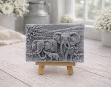 Load image into Gallery viewer, A rectangular handmade vegan soap in a charcoal grey tone, featuring a detailed relief carving of an elephant family—two adults and a small calf—walking through a textured savannah landscape, displayed on a miniature wooden easel.