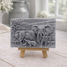Load image into Gallery viewer, A rectangular handmade vegan soap in a charcoal grey tone, featuring a detailed relief carving of an elephant family—two adults and a small calf—walking through a textured savannah landscape, displayed on a miniature wooden easel.
