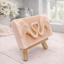 Load image into Gallery viewer, A rectangular handmade vegan soap featuring a detailed relief of two elephants facing each other with their trunks entwined to form a heart shape, displayed on a miniature wooden easel.