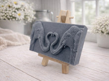 Load image into Gallery viewer, A rectangular handmade vegan soap in a slate-grey tone, featuring a detailed relief of two elephants facing each other with their trunks entwined to form a heart shape, displayed on a miniature wooden easel.