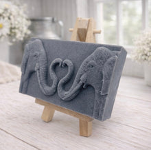 Load image into Gallery viewer, A rectangular handmade vegan soap in a slate-grey tone, featuring a detailed relief of two elephants facing each other with their trunks entwined to form a heart shape, displayed on a miniature wooden easel.