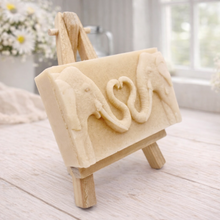 Load image into Gallery viewer, A rectangular handmade vegan soap featuring a detailed relief of two elephants facing each other with their trunks entwined to form a heart shape, displayed on a miniature wooden easel.