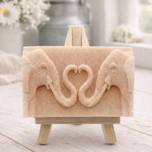 Load image into Gallery viewer, A rectangular handmade vegan soap featuring a detailed relief of two elephants facing each other with their trunks entwined to form a heart shape, displayed on a miniature wooden easel.