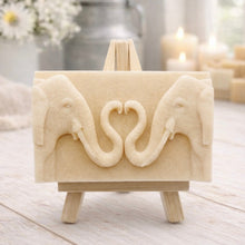 Load image into Gallery viewer, A rectangular handmade vegan soap featuring a detailed relief of two elephants facing each other with their trunks entwined to form a heart shape, displayed on a miniature wooden easel.