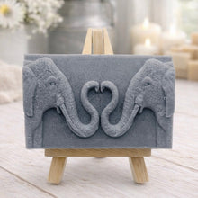 Load image into Gallery viewer, A rectangular handmade vegan soap in a slate-grey tone, featuring a detailed relief of two elephants facing each other with their trunks entwined to form a heart shape, displayed on a miniature wooden easel.