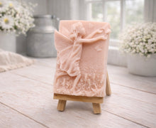 Load image into Gallery viewer, A rectangular handmade vegan soap featuring a detailed relief of a winged fairy reaching towards a butterfly in a meadow of tall grass and flowers, displayed on a miniature wooden easel