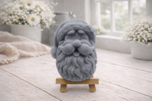 Load image into Gallery viewer, handmade vegan soap sculpted in the shape of Father Christmas's head, featuring a detailed curly beard, a jolly moustache, and a classic Santa hat, displayed on a miniature wooden easel in a bright farmhouse setting.