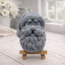Load image into Gallery viewer, handmade vegan soap sculpted in the shape of Father Christmas's head, featuring a detailed curly beard, a jolly moustache, and a classic Santa hat, displayed on a miniature wooden easel in a bright farmhouse setting.