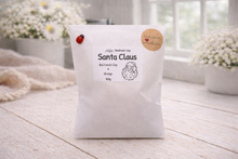 Load image into Gallery viewer, White bag with 'Santa Claus' soap label on a wooden surface with flowers in the background