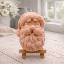 Load image into Gallery viewer, handmade vegan soap sculpted in the shape of Father Christmas's head, featuring a detailed curly beard, a jolly moustache, and a classic Santa hat, displayed on a miniature wooden easel in a bright farmhouse setting.