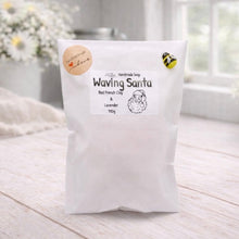 Load image into Gallery viewer, White bag labeled 'Waving Santa' on a light wooden surface with flowers in the background
