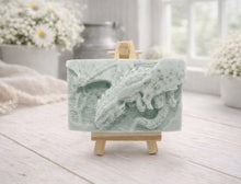 Load image into Gallery viewer, handmade soap bar featuring a detailed 3D relief carving of a dragon, displayed on a small wooden easel in a bright, rustic room with white flowers in the background.