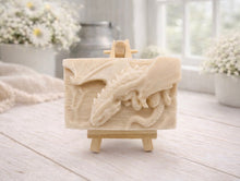 Load image into Gallery viewer, handmade soap bar featuring a detailed 3D relief carving of a dragon, displayed on a small wooden easel in a bright, rustic room with white flowers in the background.