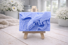 Load image into Gallery viewer, handmade soap bar featuring a detailed 3D relief carving of a dragon, displayed on a small wooden easel in a bright, rustic room with white flowers in the background.