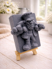 Load image into Gallery viewer, A rectangular handmade vegan soap featuring a high-relief carving of Frankenstein's monster in a classic pose with arms outstretched and a grumpy expression, displayed on a miniature wooden easel.