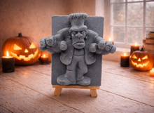 Load image into Gallery viewer, A rectangular handmade vegan soap featuring a high-relief carving of Frankenstein's monster in a classic pose with arms outstretched and a grumpy expression, displayed on a miniature wooden easel.