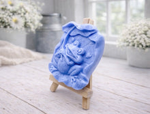 Load image into Gallery viewer, A rectangular handmade vegan soap featuring a high-relief carving of a cheerful, smiling frog sitting on a large lilypad and holding the stem of a leaf over its head like an umbrella.