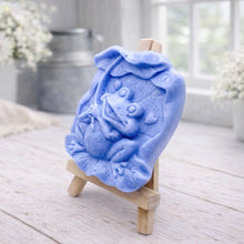 Load image into Gallery viewer, A rectangular handmade vegan soap featuring a high-relief carving of a cheerful, smiling frog sitting on a large lilypad and holding the stem of a leaf over its head like an umbrella.
