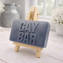 Load image into Gallery viewer, Gray soap bar with 'GAY BAR' text on a wooden stand, placed on a light wooden surface.