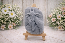 Load image into Gallery viewer, A rectangular handmade vegan soap in a natural tone, featuring a detailed relief of a mother and baby giraffe with their necks intertwined, displayed on a miniature wooden easel against a white wood-panelled background.