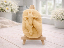 Load image into Gallery viewer, A rectangular handmade vegan soap in a natural tone, featuring a detailed relief of a mother and baby giraffe with their necks intertwined, displayed on a miniature wooden easel against a white wood-panelled background.