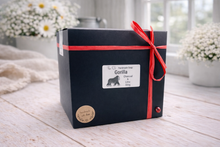 Load image into Gallery viewer, Black gift box with a red ribbon on a wooden surface with flowers in the background