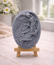 Load image into Gallery viewer, An oval-shaped handmade vegan soap, featuring an intricate relief carving of a guardian angel with large wings holding flowers, displayed on a small wooden easel.