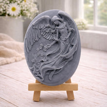 Load image into Gallery viewer, An oval-shaped handmade vegan soap, featuring an intricate relief carving of a guardian angel with large wings holding flowers, displayed on a small wooden easel.