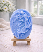Load image into Gallery viewer, An oval-shaped handmade vegan soap, featuring an intricate relief carving of a guardian angel with large wings holding flowers, displayed on a small wooden easel.