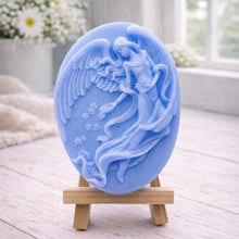 Load image into Gallery viewer, An oval-shaped handmade vegan soap, featuring an intricate relief carving of a guardian angel with large wings holding flowers, displayed on a small wooden easel.