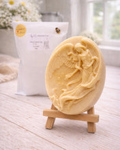 Load image into Gallery viewer, An oval-shaped handmade vegan soap, featuring an intricate relief carving of a guardian angel with large wings holding flowers, displayed on a small wooden easel.