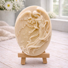 Load image into Gallery viewer, An oval-shaped handmade vegan soap, featuring an intricate relief carving of a guardian angel with large wings holding flowers, displayed on a small wooden easel.