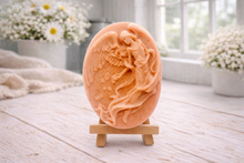 Load image into Gallery viewer, An oval-shaped handmade vegan soap, featuring an intricate relief carving of a guardian angel with large wings holding flowers, displayed on a small wooden easel.