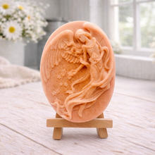 Load image into Gallery viewer, An oval-shaped handmade vegan soap, featuring an intricate relief carving of a guardian angel with large wings holding flowers, displayed on a small wooden easel.